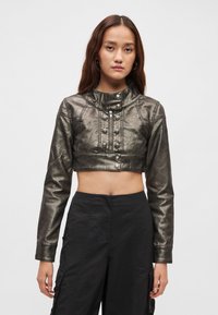 Μεταλλικό cropped μπουφάν με ψηλό γιακά, λεπτομέρειες με κουμπιά και μακριά μανίκια, κατασκευασμένο από συνθετικό δέρμα σε σκούρο γκρι. Συνδυασμένο με μαύρο παντελόνι.