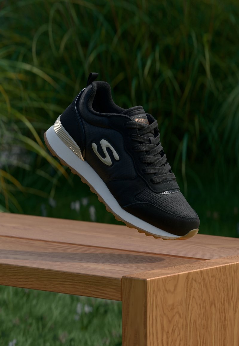 Baskets Skechers noires avec semelle blanche et détail métallique au talon, positionnées au-dessus d'un banc en bois, sur fond d'herbe verte et de feuillage.