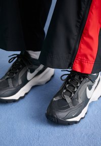 Person klädd i svarta och röda träningsbyxor samt svarta och vita Nike-sneakers står på en blå matta.