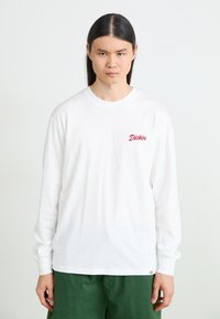 White long-sleeve t-shirt made of cotton, featuring a small red logo on the chest, round neck, and loose fit. Paired with green pants.