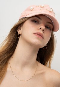 Rosa Baumwollmütze mit großen Kunstperlen und einer Kettenakzentuierung. Verfügt über einen gebogenen Schirm und eine strukturierte Form. Sichtbare goldene Creolen.