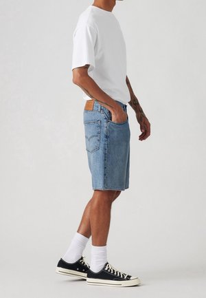 Homme portant un t-shirt blanc, un short en jean bleu clair, des chaussettes blanches et des baskets basses noires, debout de profil.