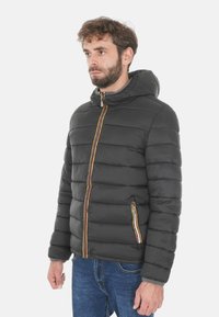 Giacca nera imbottita con cappuccio, caratterizzata da una cerniera e tasche laterali di colore arancione e giallo a contrasto. Texture liscia e design trapuntato.