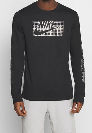 Homme portant un t-shirt Nike noir à manches longues avec un motif en quadrillage blanc et un logo sur la poitrine et les manches, associé à un short Nike gris clair.