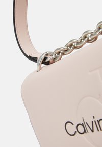 Bolso de piel color rubor con superficie texturizada, que presenta una correa de cadena plateada y un logotipo de la marca en relieve. Costuras de contraste visibles.