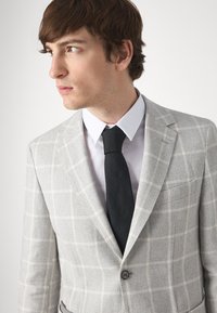 Lichtgrijze blazer van een gestructureerde stof met een wit ruitpatroon, gecombineerd met een wit overhemd en een zwarte stropdas. Opvallende enkele knoopsluiting.