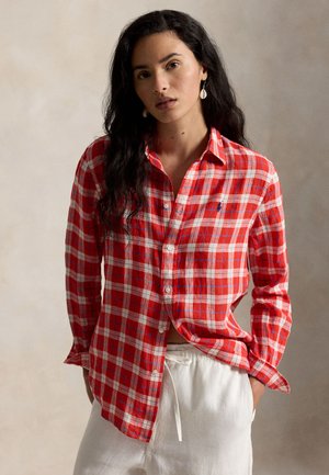 Γυναίκα με μακριά σκούρα μαλλιά φοράει κόκκινο-άσπρο πουκάμισο με κουμπιά και λευκό παντελόνι με κορδόνι, μπροστά σε απλό ουδέτερο φόντο.