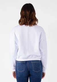 Felpa bianca con colletto a girocollo e polsini a costine, caratterizzata da una cucitura lungo il centro della parte posteriore, abbinata a jeans blu con tasche posteriori.
