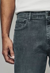 Tamno sivi traperice imaju ravnu nogavicu, dizajn s pet džepova i suptilnu teksturu. Ruka se oslanja na bočni šav.