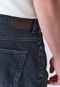 Primer plano de una persona que lleva vaqueros de mezclilla negros con un parche de cuero marrón "Denim Supply" en la cintura y el brazo derecho visible.