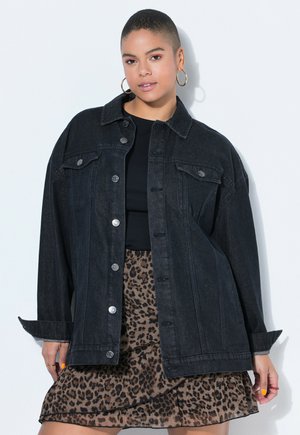 Mladá žena s krátkými vlasy, která nosí zlaté kruhové náušnice, velkou černou džínovou bundu, černý top a sukni s leopardím vzorem.