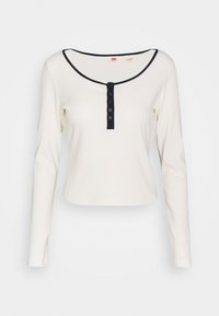 Dlouhý rukáv krémový top s žebrovanou texturou, kulatým výstřihem, námořnickým lemováním a předním zapínáním na šest černých knoflíků.