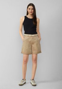 Haut sans manches noir, shorts beige à revers avec passants de ceinture, et baskets blanches avec accents gris. Le mannequin se tient les mains dans les poches.