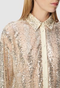 Blusa beige con paillettes e motivo intricato, materiale trasparente e abbottonatura color crema. Decorazioni scintillanti che aggiungono texture e dettagli.