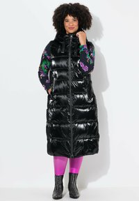 Schwarze Pufferweste mit glänzendem Finish, durchgehendem Reißverschluss, hohem Kragen und horizontaler Steppung, kombiniert mit floral gemusterten Ärmeln und pinkfarbenen Leggings.
