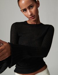 Μαύρο ριμπ μακρυμάνικο τοπ με φουσκωτά μανίκια, εφαρμοστή γραμμή και ελαφρώς διαφανή υφή. Απλός στρογγυλός λαιμός και υφασμάτινη επιφάνεια.