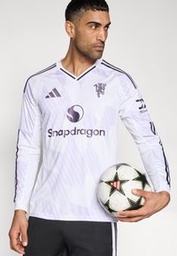 Camisola de futebol branca de manga comprida com detalhes em roxo, padrões diagonais e logótipos. O modelo segura uma bola de futebol preta e branca com estrelas.