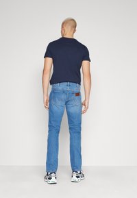 Aansluitend marineblauw T-shirt gecombineerd met lichtblauwe jeans. De jeans hebben twee achterzakken en een leren bruin label. Draagt sportieve schoenen.