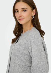 Lys grå strikket cardigan med rund halsudskæring, lange ærmer og forlommer. Teksturen ser blød og hyggelig ud.