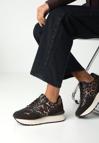 Hnědé tenisky s leopardím vzorem a kombinací texturovaných materiálů, se zlatými akcenty a polstrovanou podrážkou. Vedle jsou černé široké džíny.