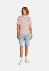 Polo de color rosa claro con un pequeño logo, combinado con pantalones cortos de mezclilla azul. El modelo lleva zapatillas blancas con calcetas rayadas.