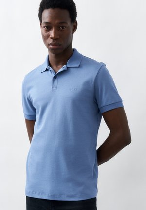 Νέος άνδρας που φοράει ανοιχτό μπλε κοντομάνικο πόλο πουκάμισο, στέκεται μπροστά σε λευκό απλό φόντο, με τα χέρια πίσω από την πλάτη του.