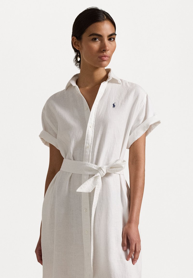 Robe en lin blanc avec ceinture, dotée d'un col, de manches courtes, de boutons à l'avant et d'un petit logo brodé sur la poitrine. Texture lisse.