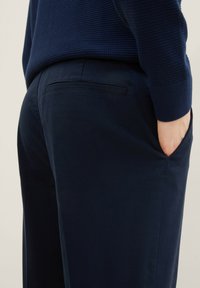 Marinblå byxor med en strukturerad väv, med en bakficka. Tyget verkar robust och slätt, vilket framhäver en avslappnad stil.