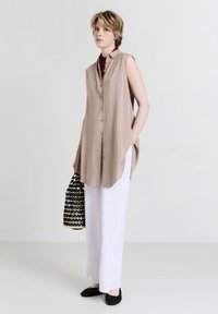 Giovane indossando una camicia senza maniche beige con bottoni, pantaloni bianchi a gamba larga, scarpe nere intrecciate, e tiene in mano una borsa intrecciata nera e beige.