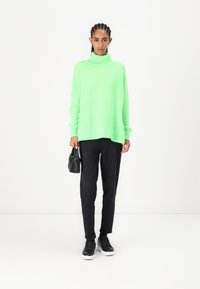 Grønn oversized turtleneck-genser med rillede mansjetter og hem, kombinert med svarte bukser og svarte sneakers. Modellen holder en liten svart veske.