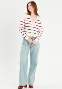 Weißer kurz geschnittener Cardigan mit bordeauxfarbenen Streifen, V-Ausschnitt und Frontbindedetail. Kombiniert mit hellblauen Schlagjeans und roten Blockabsatzschuhen.