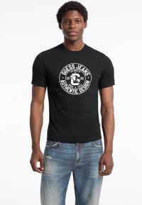 Sort bomulds t-shirt med en cirkulær hvid grafik i midten, der har teksten "Guess Jeans" og "Authentic Design". Den har rund halsudskæring og korte ærmer.