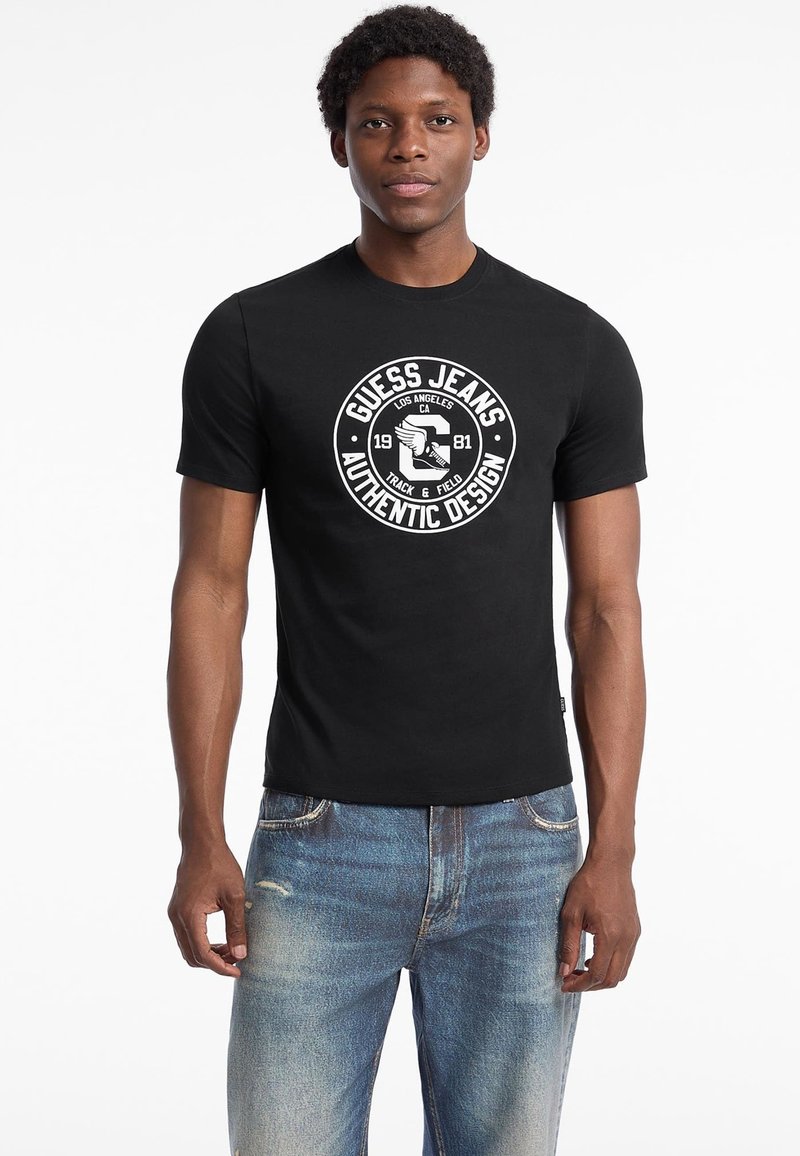Sort bomulds t-shirt med en cirkulær hvid grafik i midten, der har teksten "Guess Jeans" og "Authentic Design". Den har rund halsudskæring og korte ærmer.