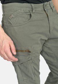 Grijze cargobroek van zachte stof met een ritszak en extra zijzakken; gestikte details langs de naden.