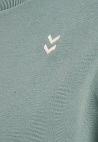 Ljusblå stickad tröja med en texturerad yta och en liten vit broderad chevronlogotyp på framsidan.