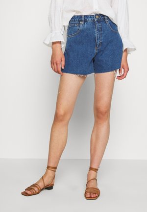 Persoon met een witte blouse, blauwe spijkershort met rafelige zomen en bruine sandalen met bandjes, staand tegen een effen achtergrond.