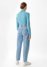 Mujer con el pelo castaño recogido en un moño, vistiendo una blusa de manga larga azul clara, vaqueros de tiro alto azul claros y zapatillas blancas, de pie de espaldas.