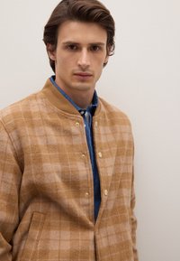 Giacca in lana a quadri marrone e beige con colletto, chiusure a scatto e tasche laterali, indossata sopra una camicia blu. Tessuto liscio.