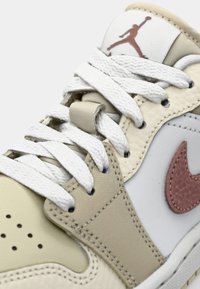 Sneaker with a cream and beige leather upper, white laces, pink textured Nike swoosh, and a brown logo on the tongue. Hexagonal perforations.
