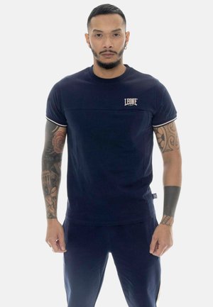 Mann mit Tattoos, trägt ein dunkelblaues Leone 1947 T-Shirt und passende Hose, steht mit Blick nach vorne vor weißem Hintergrund.