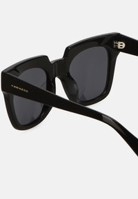 Black sunglasses with a chunky geometric frame, featuring dark lenses and a shiny finish. "Hawkers" branding displayed on the side.