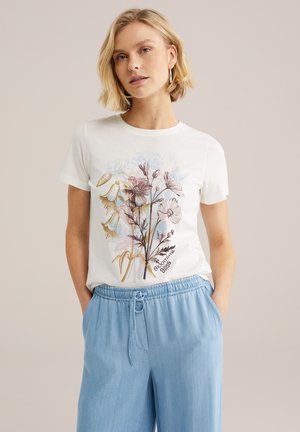Blonde vrouw met een wit T-shirt met een kleurrijke bloemenprint en de tekst 'Blooming Bliss', gecombineerd met lichtblauwe broek met trekkoord, staand met handen in de zakken.