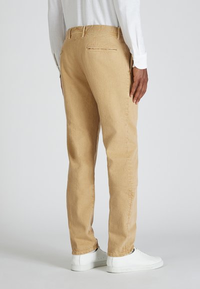 Persona que lleva pantalones rectos beige y zapatillas blancas, de pie con una mano relajada a su lado frente a un fondo sencillo.