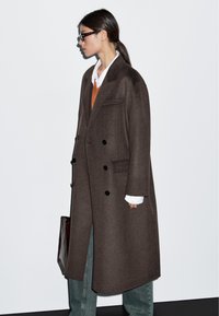 Brązowy wełniany płaszcz podwójnie zapięty z czarnymi guzikami, z klapami i bocznymi kieszeniami, noszony na pomarańczowym swetrze i białej koszuli.