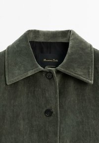 Green suede jacket featuring a notched collar, front button closure, and a smooth inner lining with a logo tag. Textured fabric detail visible.