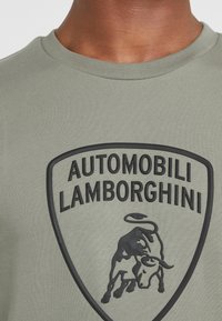Camiseta verde de tela acanalada con un logo negro elevado de Lamborghini en forma de escudo, con un diseño en relieve de un toro en el centro.