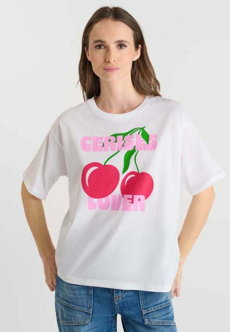 Femme portant un t-shirt blanc avec de grandes cerises rouges et du texte rose "AMOUREUSE DES CERISES", associé à un jean bleu.