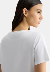 T-shirt bianco in cotone con una vestibilità rilassata, maniche corte e scollo classico a girocollo, mostrato da un'angolazione laterale.