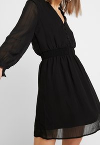 Schwarzes Kleid mit V-Ausschnitt, Knopfverschluss und langen durchsichtigen Ärmeln. Mit einem smockierten Taillenteil und einem knielangen, fließenden Rock.