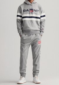 Persona che indossa un felpa grigia GANT e pantaloni della tuta coordinati con sneakers bianche, mani in tasca, su uno sfondo chiaro e semplice.